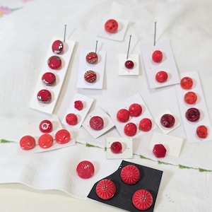 May include: A collection of various red buttons in different shapes, sizes, and textures, including smooth, patterned, and faceted designs. The buttons are mounted on small white and black rectangular cards, some secured with thin metal pins.