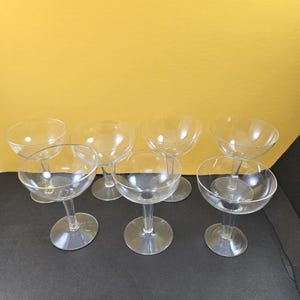 May include: A set of six clear glass coupe glasses, each featuring a wide, shallow bowl, a short, sturdy stem, and a circular base. The glasses are arranged in two rows, with three glasses in the front and three glasses positioned behind them.