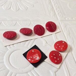 May include: A collection of seven round red buttons. Four buttons feature a split design with one smooth half and one textured half. Three buttons display a raised graphic of playing cards with white suit symbols on a glossy red surface.