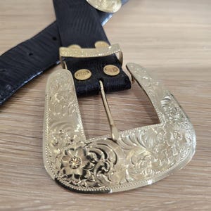 May include: A black leather belt with a gold-coloured, ornate buckle. The buckle features floral and scroll designs. The belt has a textured pattern and gold-coloured accents.