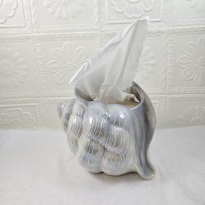 May include: A ceramic tissue holder shaped like a large conch shell with an iridescent, pearlescent finish that reflects light in shades of blue and purple. A single white facial tissue is pulled upward from the opening of the shell container.