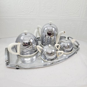 Könnte beinhalten: Ein Vintage-Teeservice aus Silber mit Teekanne, Sahnekännchen, Zuckerdose und einer kleinen Teekanne, alles auf einem Silbertablett. Das Set hat weiße Griffe und Akzente. Das Teeservice ist reflektierend.
