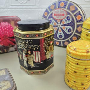May include: A selection of vintage decorative tins. The central black tin has an Asian-inspired design with figures and gold detailing. Other tins include a blue and white floral tin and yellow tins with bee patterns.
