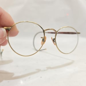 Può includere: Occhiali rotondi con una sottile montatura in metallo color oro. Le lenti sono trasparenti e i naselli sono di colore marrone chiaro. Gli occhiali sono tenuti in mano su sfondo bianco.
