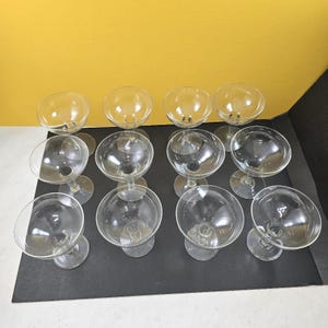May include: A set of twelve clear glass coupe glasses arranged in three rows of four. Each glass features a wide, shallow bowl, a short stem, and a circular base. The glassware is transparent and smooth, designed for serving cocktails or desserts.