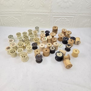 May include: A collection of numerous empty wooden and plastic sewing thread spools in various shades of beige, tan, brown, and black. The spools feature cylindrical bodies with circular ends, some with paper labels and central holes for thread.