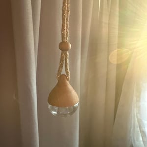 Könnte beinhalten: Ein hängender Lufterfrischer mit einem transparenten Glasboden und einer Holzoberseite. Der Lufterfrischer wird von einem goldfarbenen Seil mit einer Holzperle gehalten. Der Hintergrund ist ein weicher, weißer Vorhang.