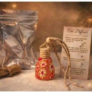 Puede incluir: Un juego de difusor de coche con un frasco de vidrio con estampado floral, una bolsa plateada y un rollo enrollado. La botella tiene una tapa de madera y una cuerda para colgar. También se incluye una tarjeta con instrucciones. Flores naranjas y blancas.