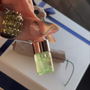 Puede incluir: Una pequeña botella de vidrio transparente llena de un líquido verde claro. La botella tiene una tapa de oro rosa y se sostiene sobre un fondo blanco y azul. La imagen está bien iluminada, resaltando el contenido de la botella.