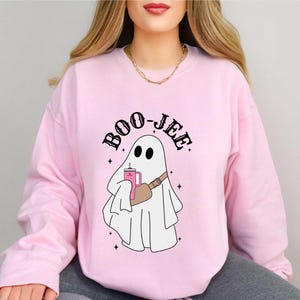 May include: Light pink sweatshirt with a graphic of a ghost holding a drink and a fanny pack. The text "BOO-JEE" is arched above the ghost. The ghost has two black eyes and is wearing a fanny pack. The sweatshirt has long sleeves.