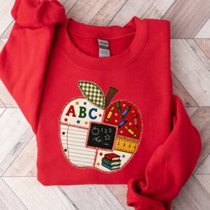 May include: A bright red crewneck sweatshirt featuring a large apple-shaped applique on the front. The apple design is divided into sections containing school-themed graphics including the letters ABC, lined paper, a chalkboard with 123, crayons, and books.
