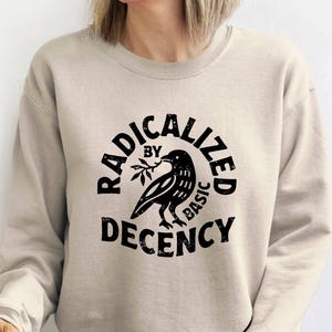 Peut inclure: Un sweat-shirt beige avec un motif noir. Le motif représente un corbeau tenant une branche, entouré des mots "RADICALIZED BY BASIC DECENCY" dans un design circulaire. Le sweat-shirt est de couleur neutre.