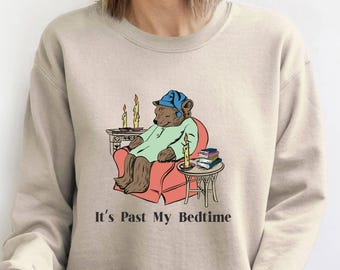 Può includere: Felpa beige con una grafica di un orso in pigiama seduto su una poltrona rossa, con candele e libri. Il testo "It's Past My Bedtime" è stampato sotto.