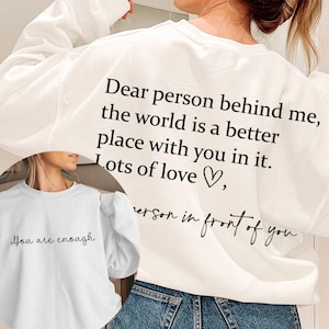 Op de afbeelding: Een wit sweatshirt met lange mouwen en zwarte tekst op de achterkant: Dear person behind me, the world is a better place with you in it. Lots of love, person in front of you. De voorkant bevat de tekst: You are enough.
