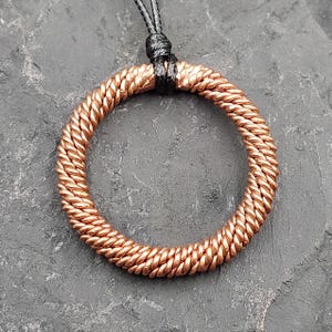 Pode incluir: Colar com pingente de cor de cobre com design circular. O pingente é feito de arame enrolado, criando uma superfície texturizada. Um cordão preto é usado para suspender o pingente. Apresentado em uma superfície texturizada cinza escuro.