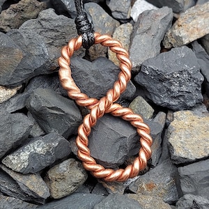 May include: A copper-colored infinity symbol pendant with a twisted rope design. The pendant is suspended from a black cord. The jewelry is displayed on a bed of dark gray rocks.