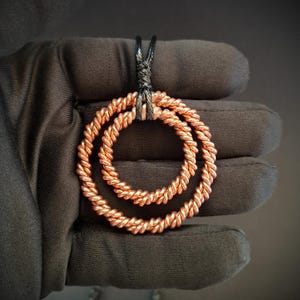 May include: A pendant featuring two concentric circles crafted from twisted copper wire. The circles are suspended from a black cord that is knotted at the top. The metallic copper material has a bright, reflective finish with a distinct rope-like texture.