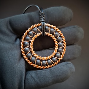 May include: A circular pendant featuring a series of smooth, dark gray spherical beads arranged in a ring, secured by a twisted copper wire frame. The pendant is attached to a black cord, creating a contrast between the metallic copper and dark beads.