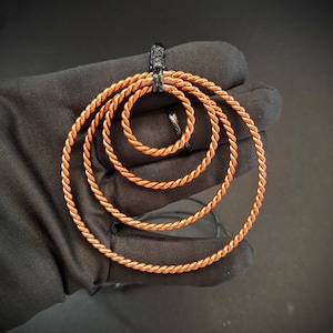 Peut inclure: Un pendentif composé de trois cercles concentriques en fil de cuivre torsadé. Les cercles sont de tailles graduées et sont reliés au sommet par une boucle en métal sombre et texturé. Le fil de cuivre présente une finition métallique brillante.