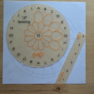 May include: A circular beige acrylic template with 15 degree spacing markings, lettered A through K, and a central flower design. The template includes the text 15 degree Spacing and Gabi. A matching beige ruler marked in inches is positioned alongside.