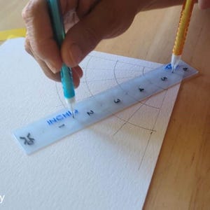 May include: A person using a clear ruler marked in inches to measure a drawing on white paper. A blue and a yellow pencil are used to draw arcs and lines. The ruler is marked with the word "INCHI".