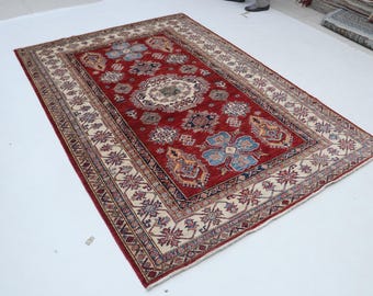 Puede incluir: Una alfombra rectangular, tejida a mano, con un campo central rojo oscuro. La alfombra presenta un detallado patrón floral en tonos azules, beige y marrón. Un borde color crema con diseños geométricos enmarca el campo central. La alfombra es probablemente de lana.