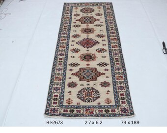 Könnte beinhalten: Ein rechteckiger beigefarbener Teppich mit einem sich wiederholenden geometrischen Muster in Rot, Blau und Schwarz. Der Teppich misst 2,7 x 6,2 Fuß, oder 79 x 189 cm. Der Teppich hat einen Rand mit floralen und geometrischen Designs.