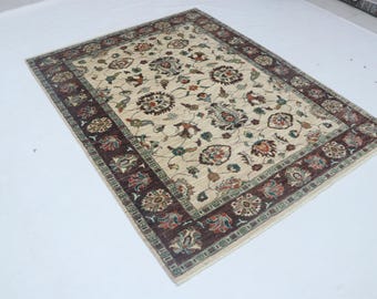 Könnte beinhalten: Rechteckiger beiger Teppich mit detailliertem Blumenmuster in Braun-, Rot- und Grüntönen. Der Teppich hat einen dunkelbraunen Rand mit sich wiederholenden Blumenmotiven. Der Teppich liegt auf einer weißen Oberfläche.