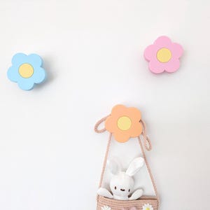 May include: Three flower-shaped wall hooks in pastel shades: blue, pink, and orange. Each hook features a yellow centre. A small woven bag with a bunny toy hangs below the orange flower hook. The hooks are mounted on a white wall.