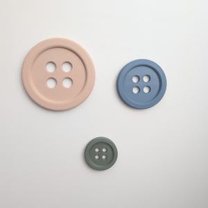May include: Three circular buttons with four holes each, arranged in a triangular formation. The largest button is light pink, the medium button is dusty blue, and the smallest button is sage green. Each button features a raised outer rim.