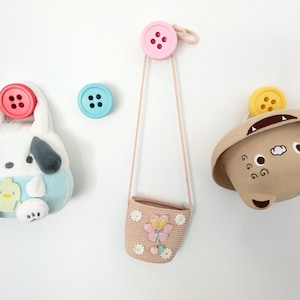 May include: A collection of whimsical wall hooks featuring button-shaped designs. The hooks hold a plush dog-shaped bag, a small woven purse with a butterfly, and a bear-shaped hat. The color palette includes pink, blue, beige, and white.