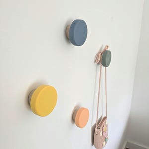 May include: A set of colourful wall hooks in yellow, blue, and peach, with a neutral base. One hook holds a small pink purse with floral detailing. These decorative hooks are mounted on a white wall.