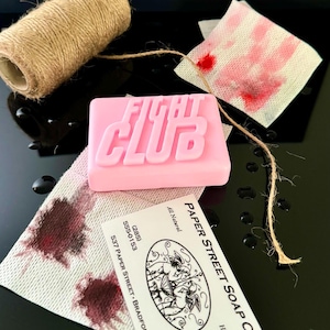 Peut inclure: Savon rose avec les mots "Fight Club" en relief sur le devant. Le savon est posé sur une serviette en papier blanche avec des taches brunes. Une carte de visite pour "Paper Street Soap Co." est visible en arrière-plan. La carte est blanche avec du texte noir et une illustration en noir et blanc de deux anges.