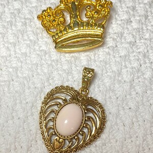 May include: Two gold-toned jewelry pieces: a crown-shaped brooch and a heart-shaped pendant. The pendant features a pale pink oval stone. Both pieces are on a white textured surface.