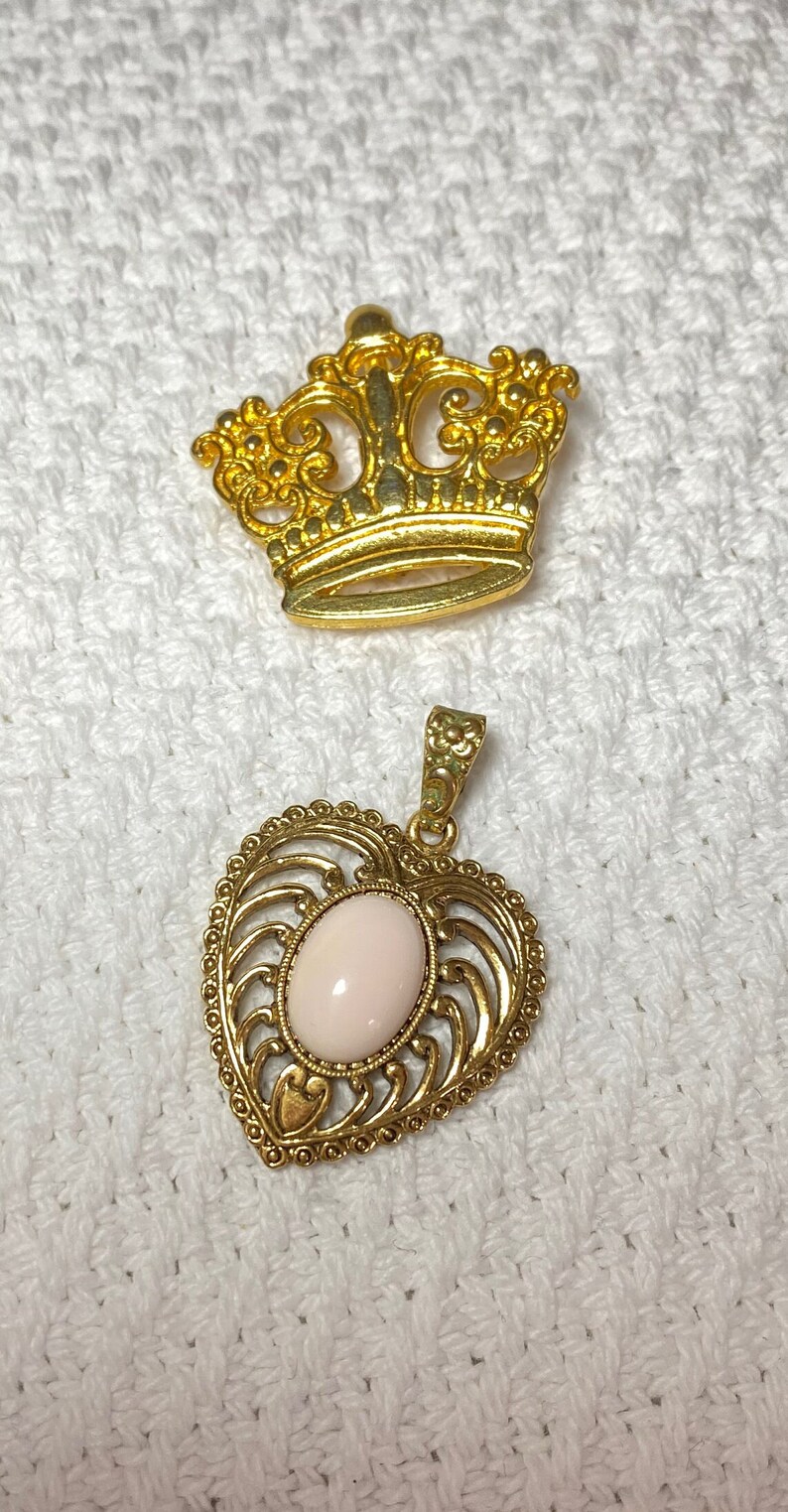 May include: Two gold-toned jewelry pieces: a crown-shaped brooch and a heart-shaped pendant. The pendant features a pale pink oval stone. Both pieces are on a white textured surface.