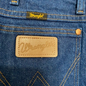 May include: Close-up of the back pocket area of blue denim Wrangler jeans. The jeans feature a brown leather patch with the Wrangler logo and a small black tag with the Wrangler logo. The stitching is a contrasting gold colour.