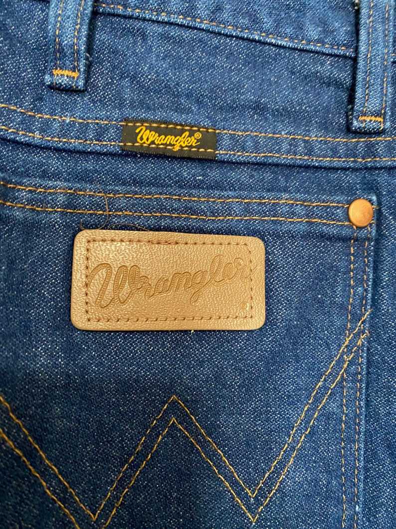 May include: Close-up of the back pocket area of blue denim Wrangler jeans. The jeans feature a brown leather patch with the Wrangler logo and a small black tag with the Wrangler logo. The stitching is a contrasting gold colour.
