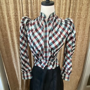 May include: A vintage-style blouse with a high collar and long sleeves. The blouse features a checkered pattern in shades of burgundy, light blue, and white. The sleeves have a puffed design at the shoulders. The bottom half of the blouse is paired with a black skirt.