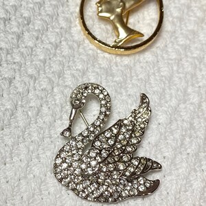 May include: Two decorative brooches. One is a gold-toned circular brooch with a profile of a woman. The other is a silver-toned swan brooch encrusted with clear crystals. Both are on a white textured surface.
