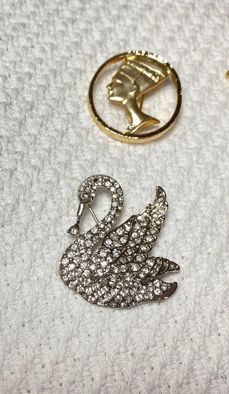May include: Two decorative brooches. One is a gold-toned circular brooch with a profile of a woman. The other is a silver-toned swan brooch encrusted with clear crystals. Both are on a white textured surface.