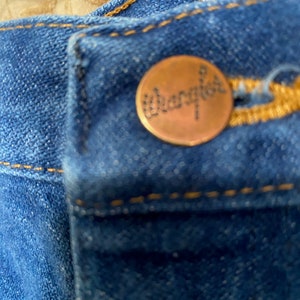 May include: Close-up of blue denim jeans with a copper-coloured button. The button is embossed with the brand name "Wrangler" in a cursive font. The jeans have yellow stitching.