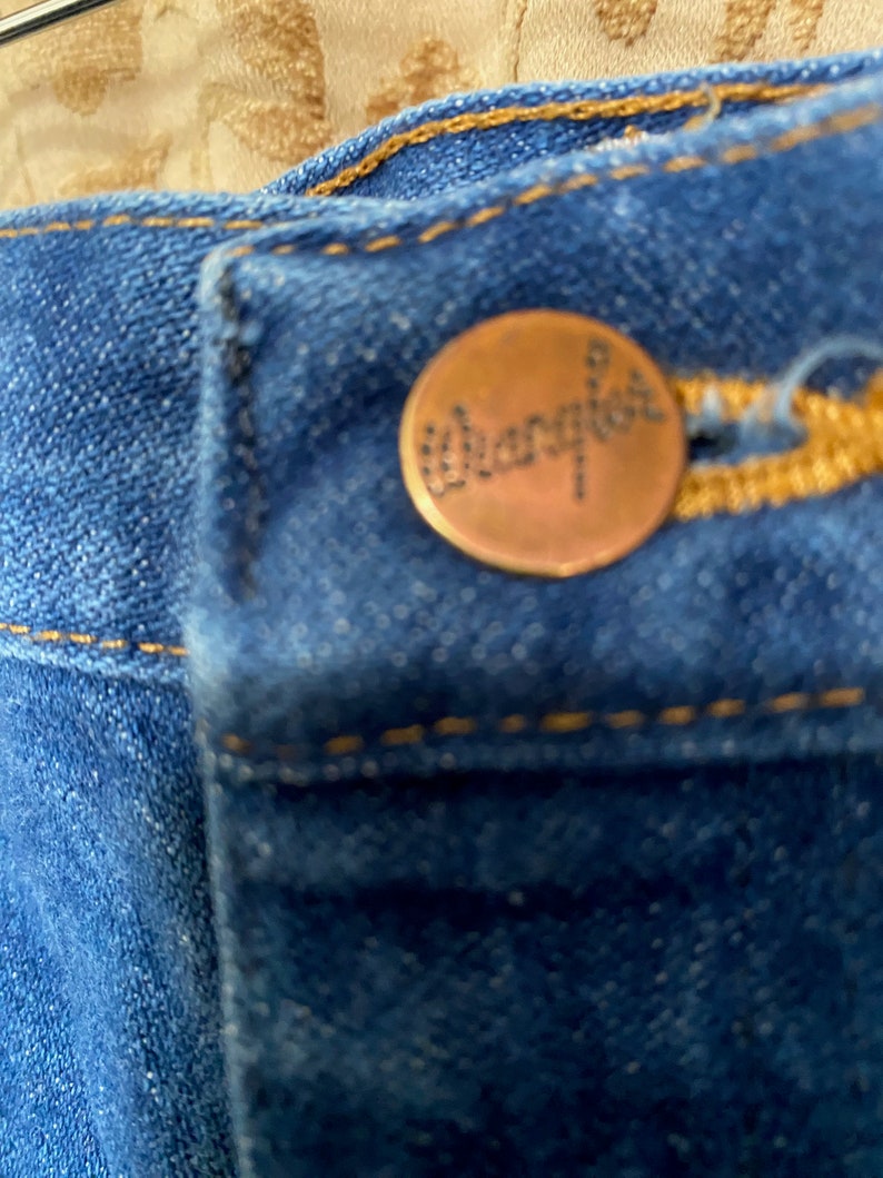 May include: Close-up of blue denim jeans with a copper-coloured button. The button is embossed with the brand name "Wrangler" in a cursive font. The jeans have yellow stitching.