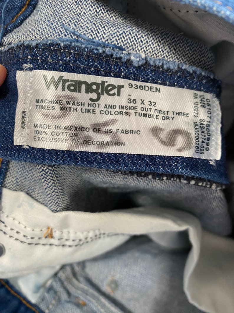 May include: Close-up of a blue denim Wrangler jeans label. The label reads "Wrangler 936DEN 36 X 32" and includes washing instructions. The jeans are made of 100% cotton and made in Mexico of US fabric.