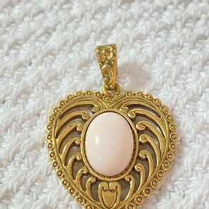 May include: A gold-colored heart-shaped pendant with an oval, light pink stone in the center. The pendant has an ornate, openwork design and a decorative bail. The pendant is likely made of metal.