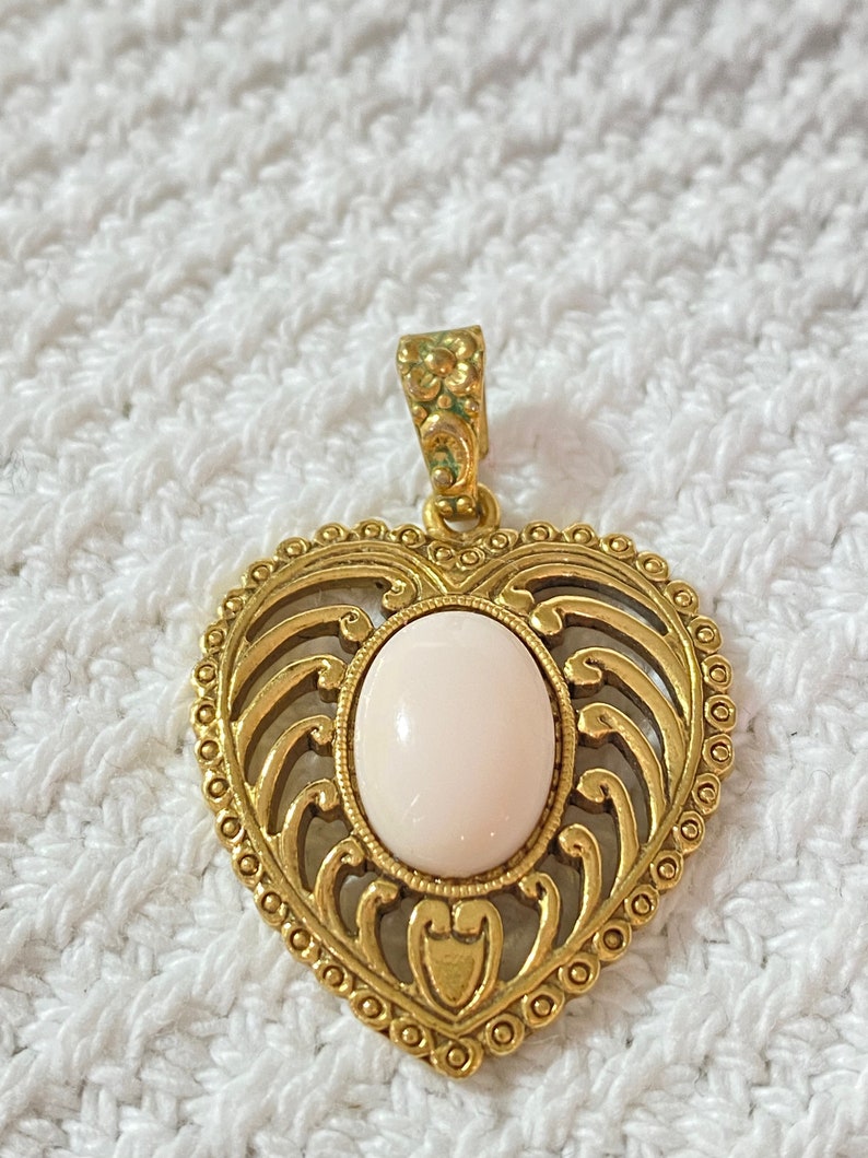 May include: A gold-colored heart-shaped pendant with an oval, light pink stone in the center. The pendant has an ornate, openwork design and a decorative bail. The pendant is likely made of metal.