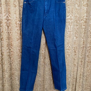 May include: A pair of blue denim jeans hanging on a clothes hanger. The jeans feature a classic five-pocket design with copper-coloured rivets and a button closure. The denim appears to be of medium weight, and the jeans have a straight-leg cut.