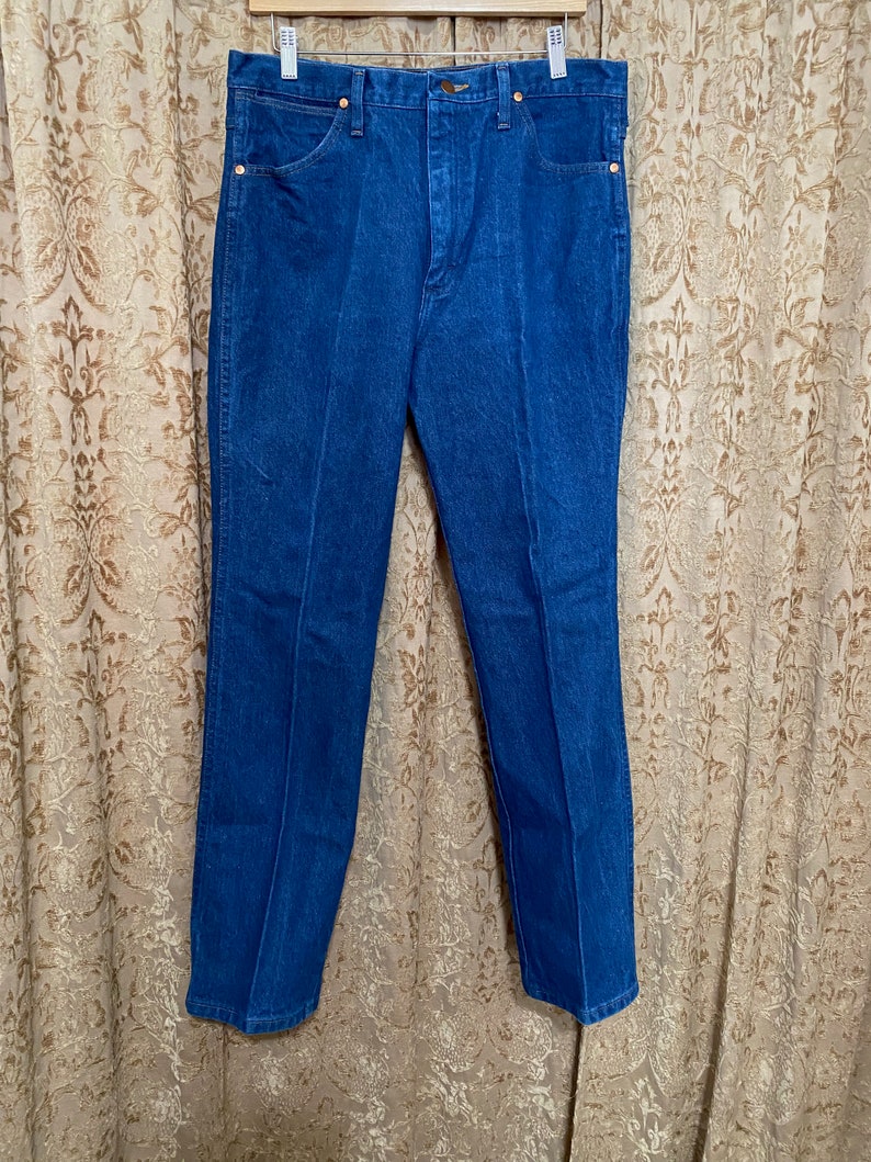 May include: A pair of blue denim jeans hanging on a clothes hanger. The jeans feature a classic five-pocket design with copper-coloured rivets and a button closure. The denim appears to be of medium weight, and the jeans have a straight-leg cut.