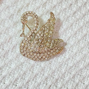 May include: A gold-toned swan brooch encrusted with clear rhinestones. The swan is depicted with its neck curved and wings spread, creating a sparkling, elegant accessory. The brooch is set against a white, textured background.