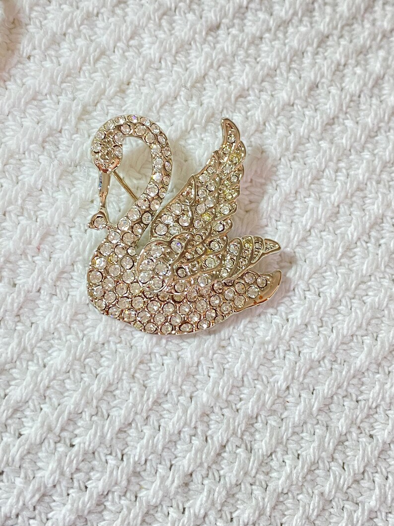 May include: A gold-toned swan brooch encrusted with clear rhinestones. The swan is depicted with its neck curved and wings spread, creating a sparkling, elegant accessory. The brooch is set against a white, textured background.