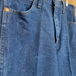 May include: Close-up of a pair of dark blue denim jeans. The jeans have copper-coloured rivets and stitching. The jeans are hanging on a wooden hanger.